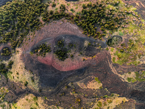 Aerial view of the volcanic landscape of Mount Etna featuring dark lava flows, red volcanic soil, and patches of green pine forest in Catania, Sicily, Italy.