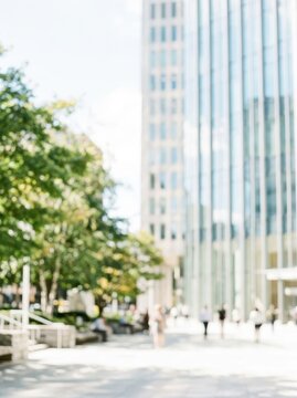 Blurred Modern City Street with Office Buildings, Green Trees, and People Walking on a Sunny Day