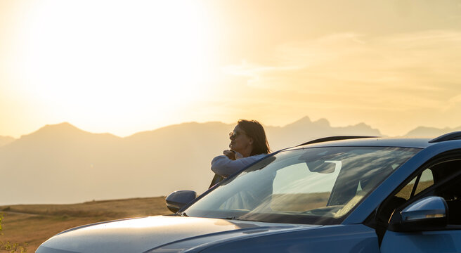 Side view of mature woman in sunglasses leaning on car door, admiring mountain sunset during road trip