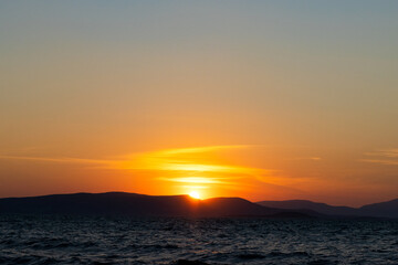 Obraz premium A dramatic sunset over rippling ocean water with the sun partially hidden behind dark mountain silhouettes, casting a vibrant orange and yellow glow across the sky.