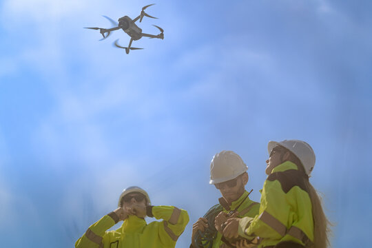 Wind technicians squint upward into the haze with hands near their faces while the inspection drone shrinks to a dark speck against the pale afternoon sky, steady green generation
