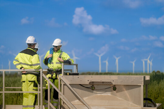 Two men in yellow high visibility coveralls lean on the handrail of a raised platform and study the working wind farm spread out across the flat green landscape