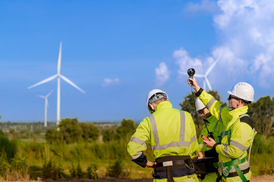 Wind speed reading beside a service road, an anemometer raised into the air by one technician while his colleague watches the meter display closely on the open Thai plain