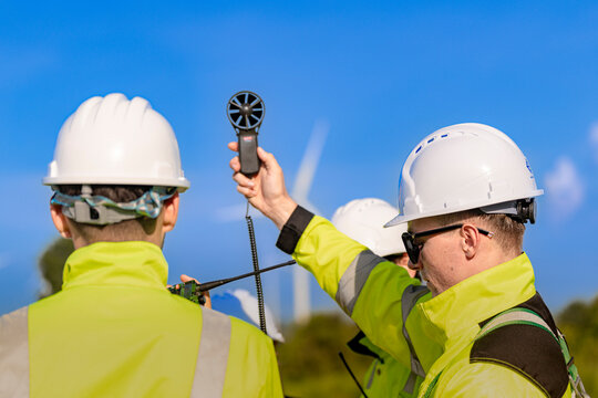 Handheld anemometer held aloft between two workers in yellow coveralls, the impeller spinning against a deep blue sky just above their white hard hats in the sun