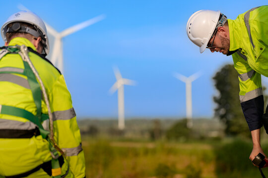 Back view of a harnessed technician standing tall over a crouching colleague, green webbing crossing his yellow jacket as turbine towers blur into the treeline on a working wind power site