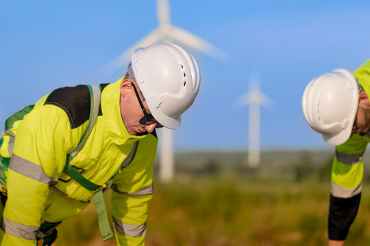 Side view of a rope access technician bending deep at the waist to lift a heavy coil of climbing line out of the grass, turbine towers blurred across the horizon