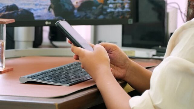 Wide shot of a woman using a smartphone at an office workstation, with a computer keyboard and monitor in the background, illustrating mobile communication, technology and modern office work.