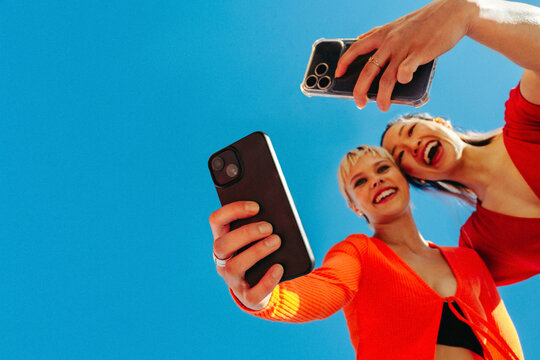 Joyful friends separately taking selfies against a clear blue sky