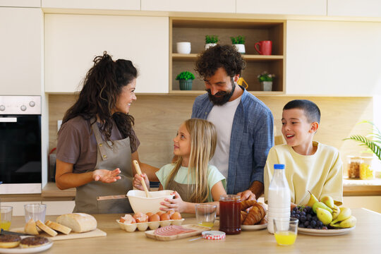 Happy family, parents and two children, enjoying a relaxed morning at home, preparing a healthy and delicious breakfast with fresh ingredients in their bright, contemporary kitchen
