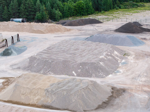 Aerial view of an asphalt concrete production plant featuring piles of gravel, sand, and aggregate materials in an open yard near a forest edge in Tume Parish, Tukums Municipality, Latvia.
