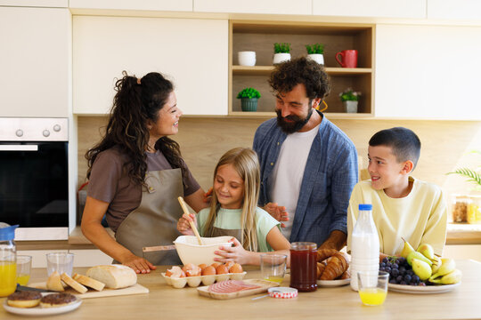 Family enjoying a morning in the kitchen, a young daughter enthusiastically mixing ingredients in a bowl while her parents and brother watch, all smiling and sharing a moment of domestic joy