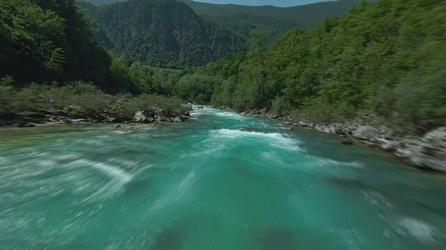 Fast moving aerial drone view flying over clear turquoise mountain river water flowing between rocky shores and lush green forest.