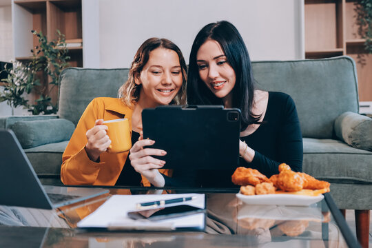 Two young female students using a laptop while sitting on a sofa at home. Student life concept with studying, teamwork, and online learning.