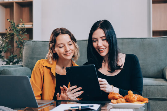 Two young female students using a laptop while sitting on a sofa at home. Student life concept with studying, teamwork, and online learning.