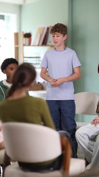 Teen group sit in circle and listen to Caucasian boy classmate speaking, teacher watching. Acting masters courses, stage speech courses, public speaking courses 