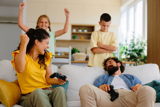 Family members, mother and daughter cheering on a victory while playing video games with controllers in the living room, dad resting on the sofa and son looking displeased