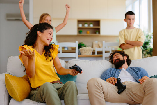 Happy family members playing video games in the living room, with a mother celebrating a win and her daughter cheering, while the father rests and the son watches the competition