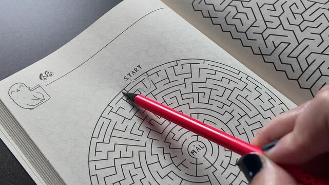 Hand solving a printed maze with a red pencil in a puzzle book. Close-up of a classic labyrinth activity representing problem solving, concentration, logical thinking, brain training and relaxing ente