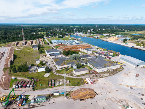 Aerial view of the industrial timber processing facility with large log piles, warehouses, and machinery situated along the canal in Mersrags, Talsi Municipality, Latvia.