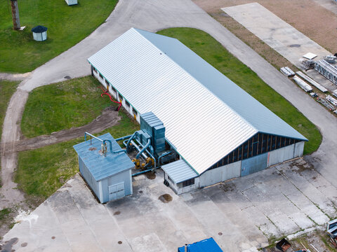 Aerial view of an industrial facility with a large metal-roofed building and external ventilation machinery situated on a concrete lot in Mersraga pagasts, Talsu novads, Latvia.