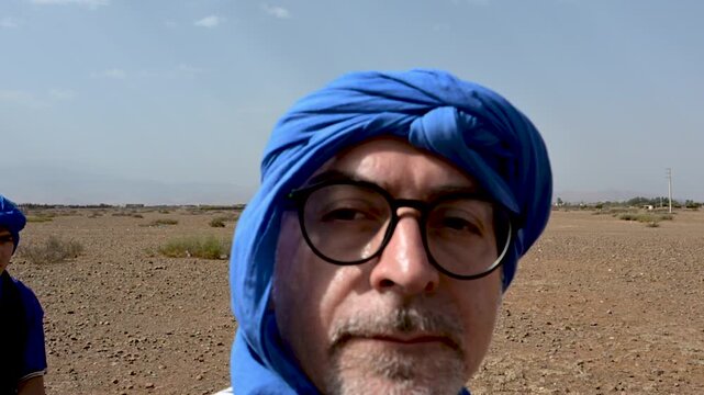 Marrakech, morocco. August 9, 2026. Selfie perspective of a mature male tourist riding a dromedary camel. A local berber guide is in the background, crossing the rocky desert