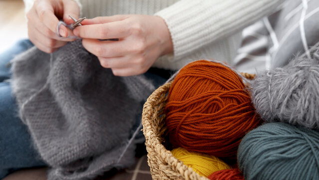 Woman wearing a white sweater and working with knitting needles beside a basket filled with colorful yarn