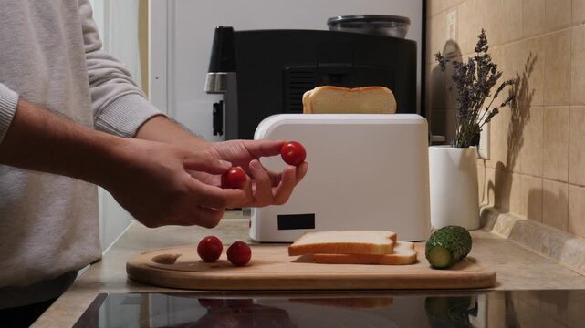 Person's hands holding fresh cherry tomatoes, preparing a healthy breakfast or snack in a kitchen, with toast in a toaster and cucumber on a wooden cutting board