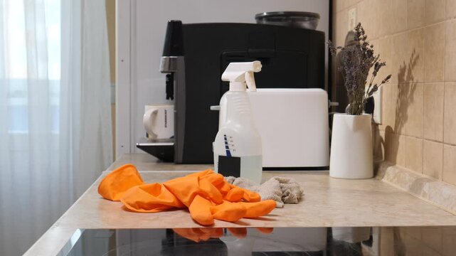 Person wearing protective orange gloves, preparing for cleaning tasks in a home kitchen, using a spray bottle and cloth to sanitize surfaces and maintain hygiene