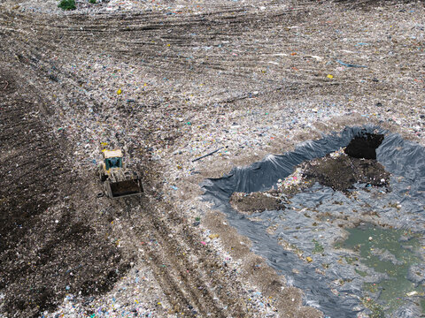 Aerial view of a bulldozer working at a municipal solid waste landfill with piles of refuse and a lined drainage pond in Laidzes pagasts, Talsu novads, Latvia.