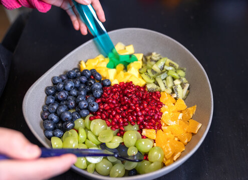 Woman mixes kiwi, oranges, pineapple, blueberries, grapes, and pomegranate seeds in a bowl to make a fruit salad. Sweet food, healthy eating, organic, ripe, preparing a salad, ready to eat.