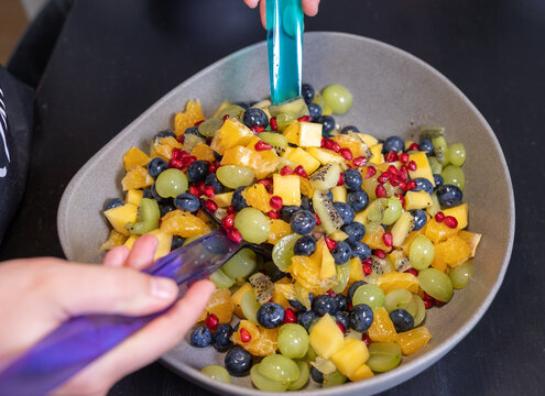 Woman mixes kiwi, oranges, pineapple, blueberries, grapes, and pomegranate seeds in a bowl to make a fruit salad. Sweet food, healthy eating, organic, ripe, preparing a salad, ready to eat.