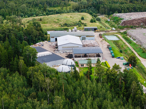 Aerial view of a municipal solid waste landfill facility with industrial buildings, storage tanks, and waste management infrastructure surrounded by forest in Laidzes pagasts, Talsu novads, Latvia.