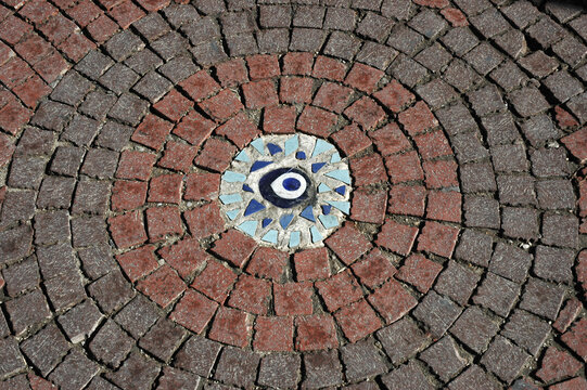 A top view close up of circular red and gray cobblestone pavement featuring a central ceramic mosaic depicting a blue and white evil eye protection symbol.