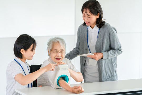 血圧計を使い血圧測定をするシニア女性と記録をとる看護師,介護士の女性スタッフ