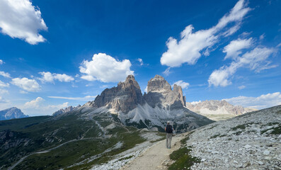 Naklejka premium hiker trekking mountain path behind rocky terrain with blue sky and white clouds, adventure third-person perspective natural light clean background editorial use.