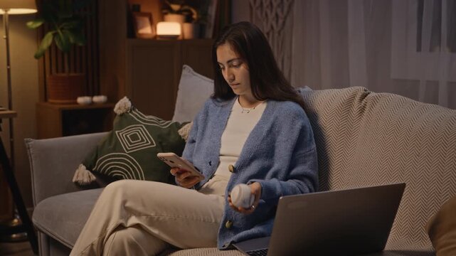 Caucasian young adult woman sitting on sofa at night, looking at smartphone while tossing stress ball in hand, laptop open nearby. Clip is ideal for marketing campaigns focusing on stress relief
