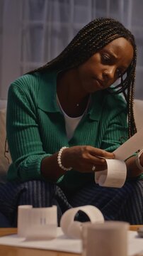 Vertical video of African American young adult woman sitting on sofa and analyzing paper receipts with stressed expression. Clip is ideal for financial planning and debt management commercials