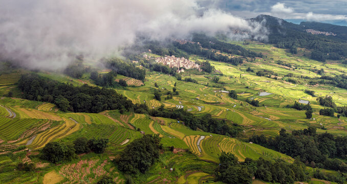 Aerial view of Yuanyang rice terraces with lush green fields, rolling hills, a village, and clouds over the mountains in Honghe Hani and Yi Autonomous Prefecture, Yunnan, China.