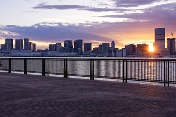 夕日のもと対岸に高層ビル群が広がる海沿い広場の風景（横構図） © Riz