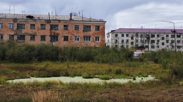 Two weathered apartment buildings stand behind overgrown vegetation and a stagnant, algae-covered pond under a cloudy sky, with no visible movement or change throughout the sequence.