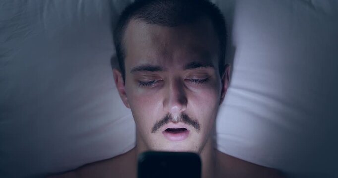 Top Down Portrait of Young Caucasian Man Lying in Bed at Night Using Smartphone in Dark
