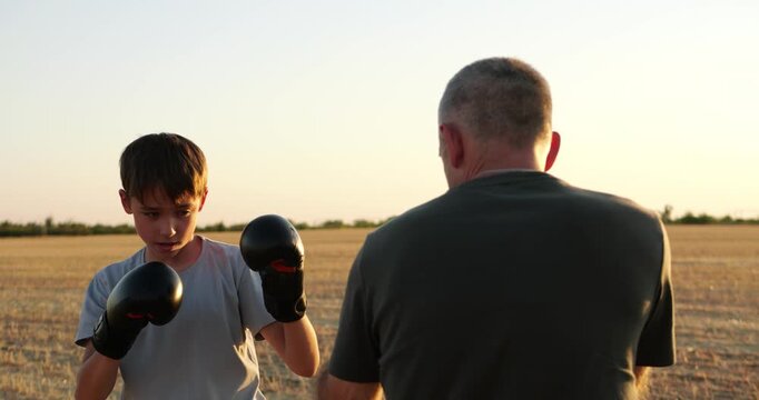 Dad and kid share boxing training in field at sunset. Young boy trains with gloves while father coaches and holds mitts. Outdoor fitness builds confidence, discipline, and family connection.