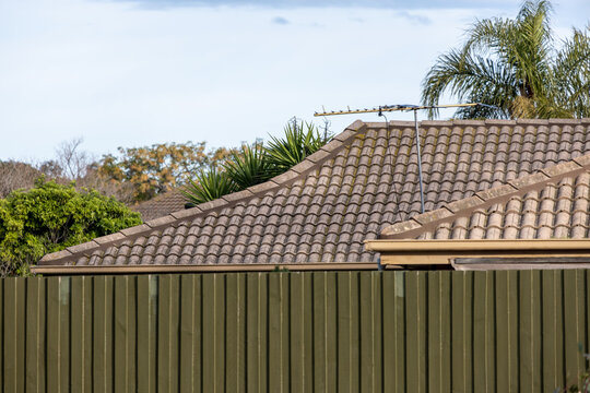 A modest suburban home's terracotta tiled roof with a TV antenna, seen above a weathered wooden fence in Melbourne, Victoria Australia.