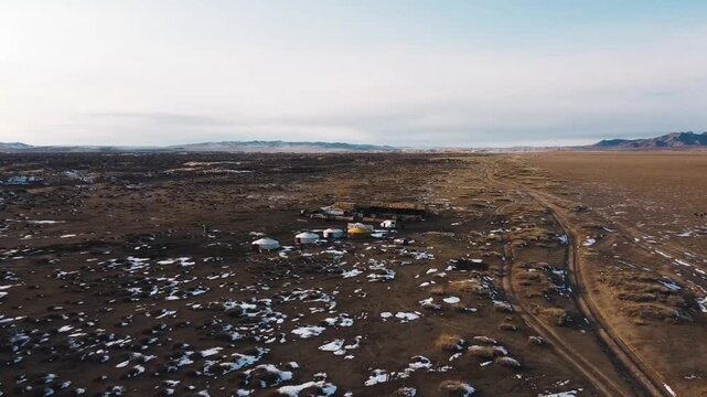 Aerial view of yurts settlement and next road surrounded by vast steppe grassland with patches of snow and overcast sky, with tracks and people in Mini Gobi, Mongolia
