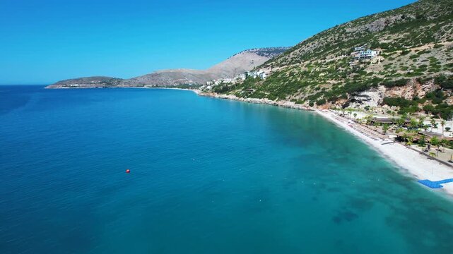 Aerial drone footage over tranquil blue waters toward a scenic beach with lush green mountains and a clear sky.