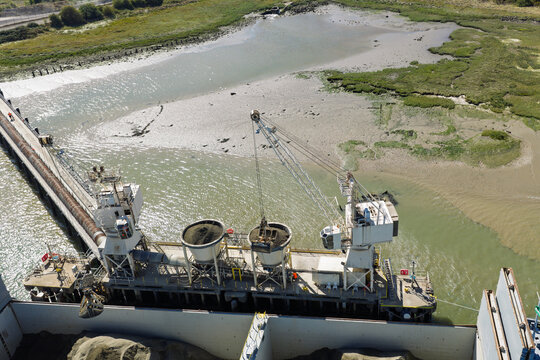 Industrial Crane Unloading Sand Cargo From a Vessel at a River Terminal During a Bright Sunny Day