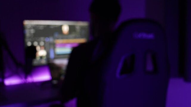 Video editor at desktop PC sits down and begins cutting a timeline on editing software, blurred static camera view with purple neon lighting, gaming chair and monitor in home studio.