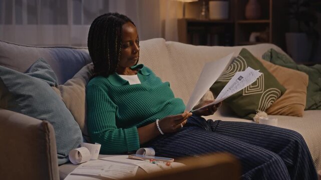 African American young adult woman sitting on sofa at night, examining past due bills and checking receipts. Clip is suitable for banking, debt relief, and financial literacy marketing campaigns
