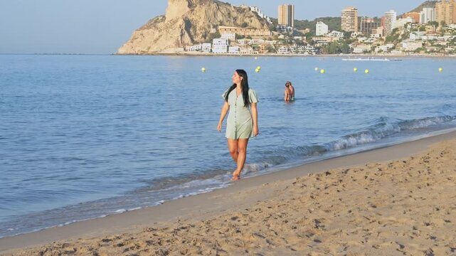 A person walks along the sandy beach in Altea, Spain. The water is clear and blue as other people enjoy the beach. Buildings and hills are in the background.