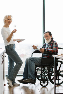 An elderly couple shares a meal, with the man seated in a wheelchair and the woman standing beside him